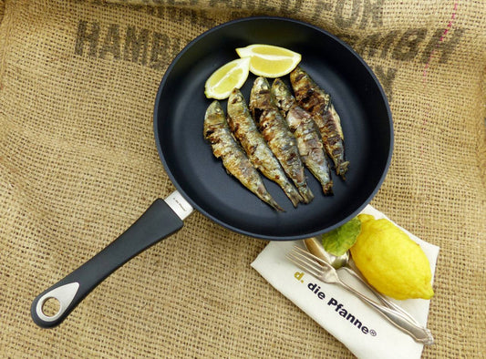 Flache Aluguss-Pfanne mit abnehmbarem Stiel und gegrillten Fischen, gelbe Zitrone und Besteck auf Juteunterlage.