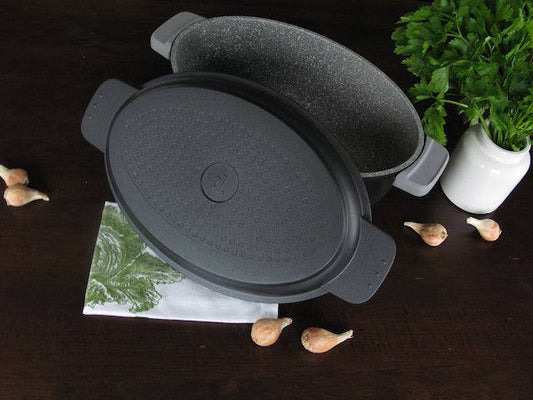 Edition 5 - der vielseitige ovale Multi-Bräter aus Aluguss mit Antihaftbeschichtung und Multifunktionsdeckel auf dunklem Tisch, umgeben von Kräutern und Knoblauch.