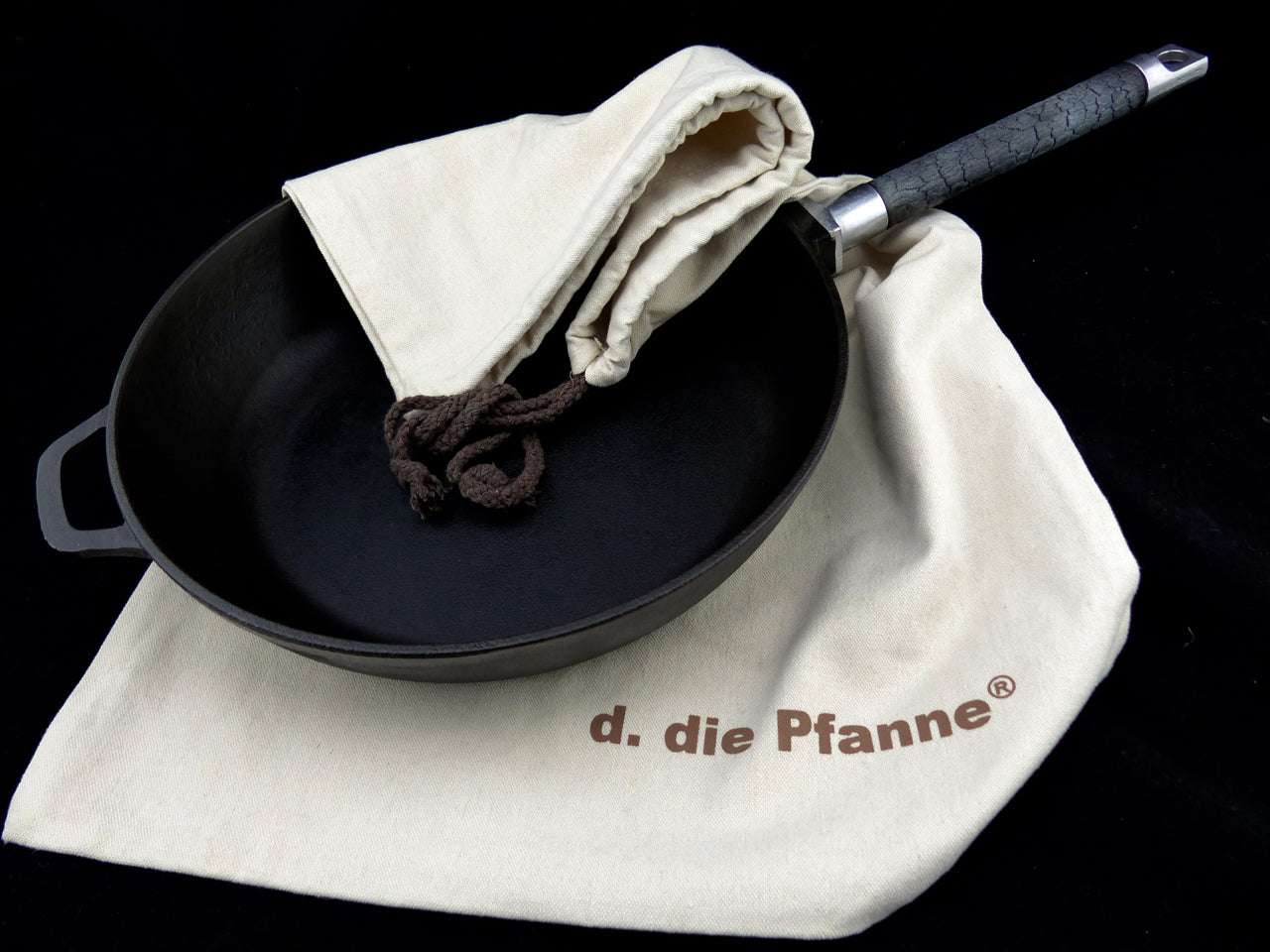 Gusseisen-Bratpfanne PUREiron mit abnehmbarem FlameWood-Stiel, liegend auf naturfarbenem Textilsäckchen von d. die Pfanne®.