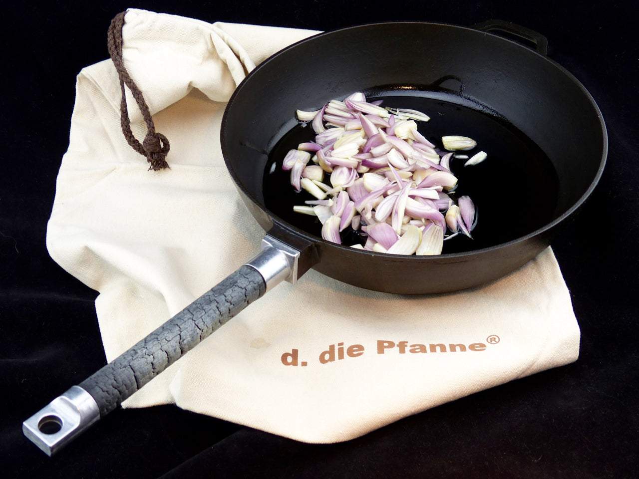 Gusseisenpfanne PUREiron mit abnehmbarem Stiel aus FlameWood, auf Stoffbeutel liegend, gefüllt mit Zwiebelstücken.