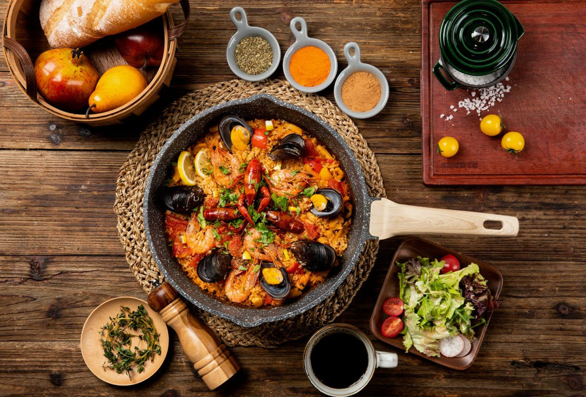 Hochrandpfanne der Aluguss-Serie Woody von Carote® mit Meeresfrüchte-Paella, umgeben von Gewürzen und Salat auf Holzuntergrund