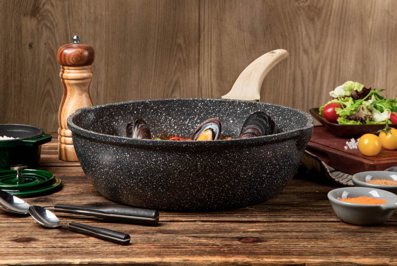 Hochrandpfanne der Aluguss-Serie Woody von Carote mit Soft-Touch-Holzeffekt-Stiel, gefüllt mit Muscheln, auf einem rustikalen Tisch.
