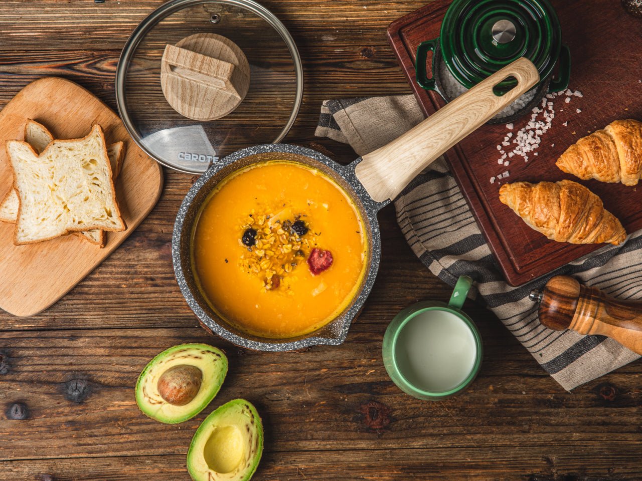 Stielkasserolle aus Aluguss mit Holzgriff und Glasdeckel, gefüllt mit Suppe, umgeben von Brot, Avocado und Croissants auf Holztisch.