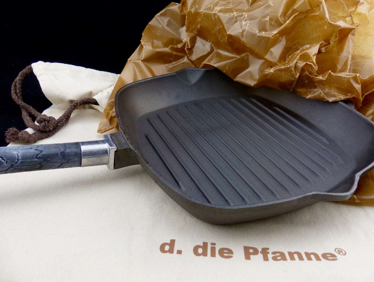 Grillpfanne PUREiron aus Gusseisen mit abnehmbarem FlameWood-Stiel, eingewickelt in nachhaltigem Paraffinpapier auf einem Textilsäckchen.