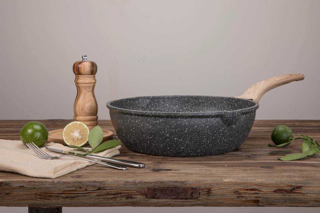 Hochrandpfanne Woody aus Aluguss mit Soft-Touch-Holzeffekt-Stiel und Granitstein-Beschichtung auf Holztisch, dekoriert mit Limette und Pfeffermühle.