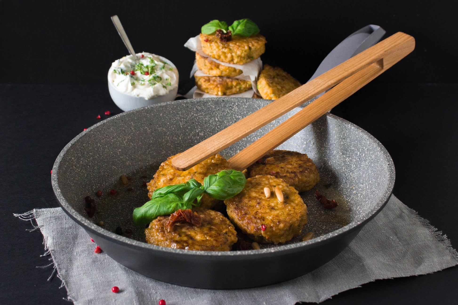 Universalpfanne aus Aluguss Edition 1 von die Pfanne mit Bratlingen und Holzzange sowie Dekoration (Basilikumzweig, Frischkäsedip und Küchentuch).. 