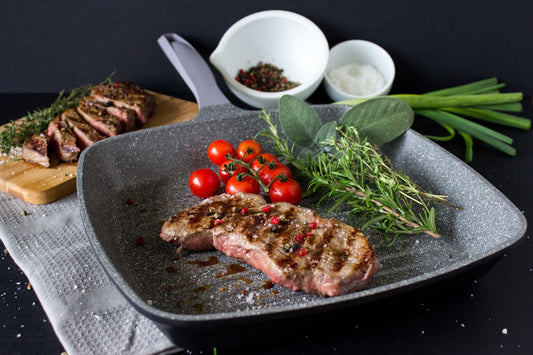 Square grey cast aluminum grill pan with grilled steak, cherry tomatoes, and fresh herbs on a dark background